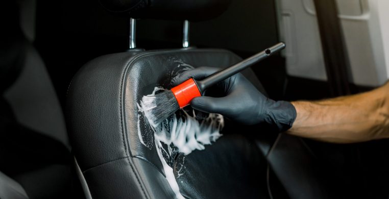Close up cropped image of washing car interior and seat by a soft brush with foam. Young male worker in black protective gloves cleaning car seat with foam and brush.